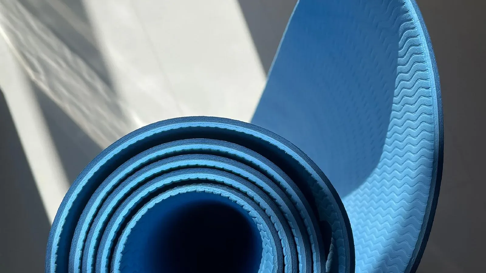 Tấm thảm yoga màu xanh cuộn tròn đặt trên sàn sáng, có nắng chiếu qua cửa sổ